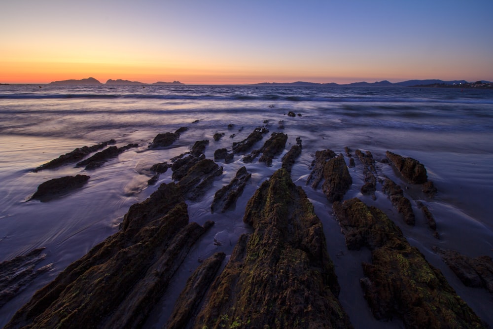 the sun is setting over the ocean with rocks in the foreground