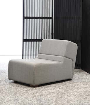 A modern, minimalistic lounge chair with a sleek grey fabric upholstery sits on a textured carpet. The backdrop features a vertical slatted screen, enhancing the contemporary aesthetic.