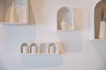 A minimalist wall display features small niches containing beige vases and candles. The decor is simple and elegant with subtle shadows cast on the light-colored wall.
