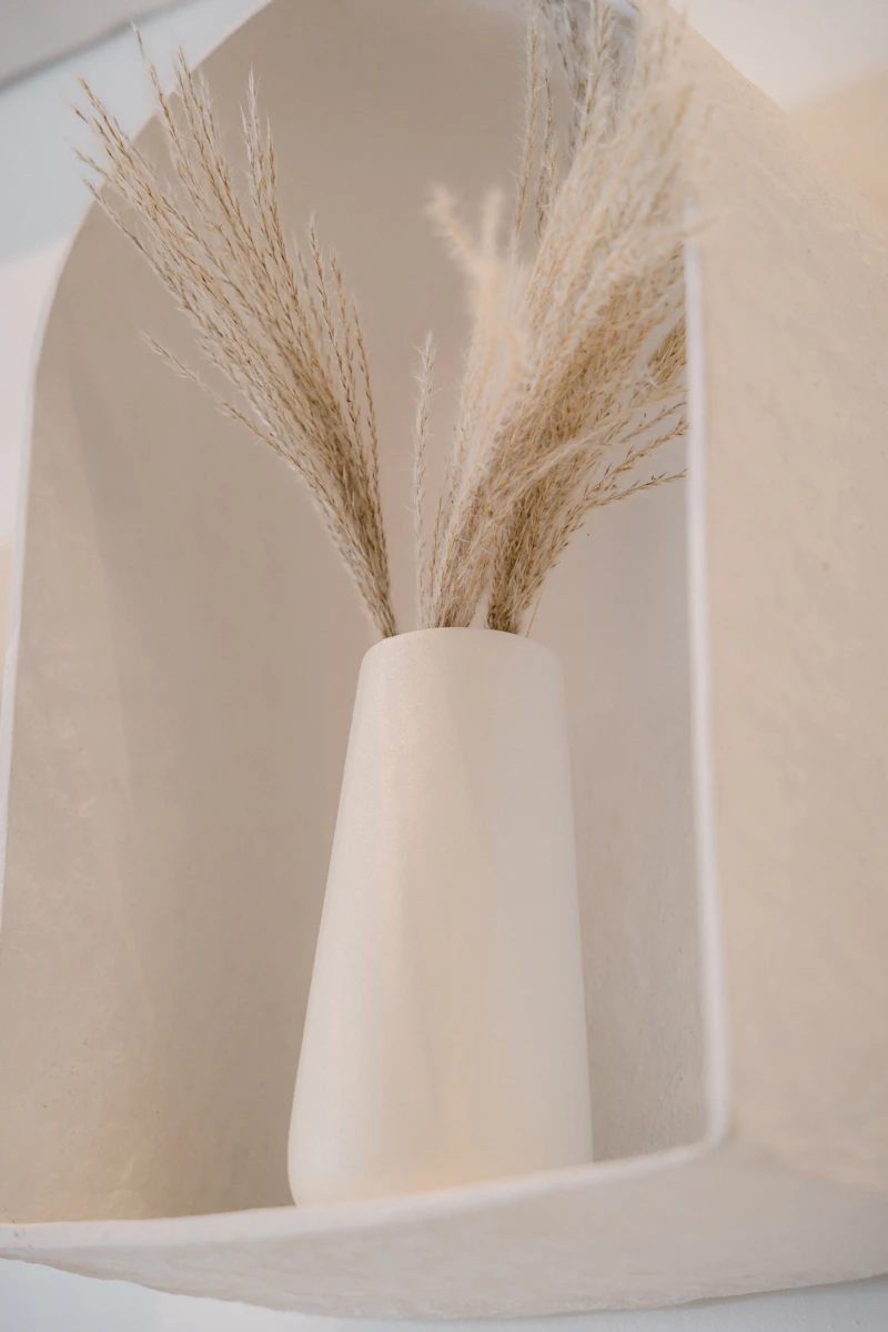 A white ceramic vase holding dried grasses on a quiet shelf