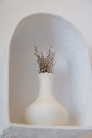 A serene corner with a single ceramic vase holding dried grasses against a pale wall.