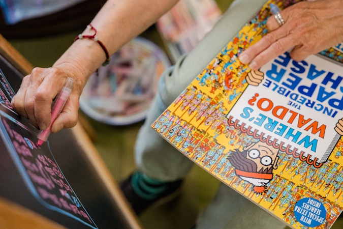 A person is holding a 'Where's Waldo?' book while writing or drawing on a blackboard with a pink marker. The cover of the book features cartoon characters and vibrant colors.