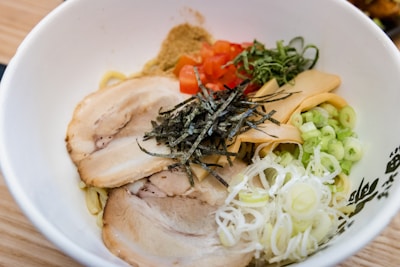 A bowl of ramen features slices of pork, chopped green onions, shredded seaweed, strips of bamboo shoots, diced red peppers, and a sprinkle of seasoning. The ingredients are beautifully arranged on top of a bed of noodles.