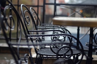 Iron chairs with intricate scrollwork surround a round table in an outdoor setting. The chairs and table appear slightly wet, suggesting recent rainfall, with some raindrops still visible on the surfaces. The background is softly blurred, hinting at a calm and possibly cool environment.