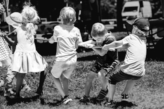 A group of children is engaged in a game of tug of war on a grassy field. The image is in black and white, highlighting the determination and teamwork of the kids. Each child has a firm grasp on the rope, pulling with effort and focus. They are casually dressed, and the background shows trees and some parked vehicles.