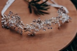 A delicate headband decorated with intricate floral and pearl designs is placed on a wooden surface. The headband features small white flowers, pearls, and metallic elements, giving it an elegant and ornamental appearance.