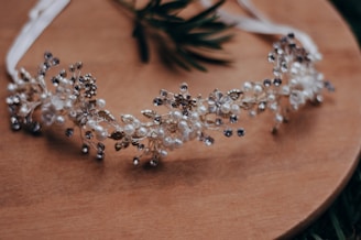 A delicate headband decorated with intricate floral and pearl designs is placed on a wooden surface. The headband features small white flowers, pearls, and metallic elements, giving it an elegant and ornamental appearance.