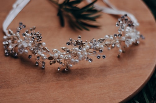 A delicate headband decorated with intricate floral and pearl designs is placed on a wooden surface. The headband features small white flowers, pearls, and metallic elements, giving it an elegant and ornamental appearance.