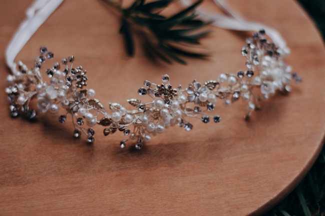 A delicate headband decorated with intricate floral and pearl designs is placed on a wooden surface. The headband features small white flowers, pearls, and metallic elements, giving it an elegant and ornamental appearance.