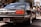 A polished, black vintage Nissan Cedric car is parked on a street with several people gathered nearby. The car has a retro design with boxy edges, reflecting a classic style. A Japanese license plate is visible, and the background includes trees and architectural features.