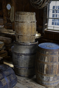 A rustic interior with several wooden barrels of varying sizes, arranged on a wooden floor. The setting includes a wall with wooden planks, an old clock, ropes, and stacks of wood. Natural light filters through a multi-paned window, creating a vintage, workshop-like atmosphere.