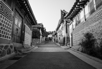 A black and white photograph of a traditional street with Korean hanok houses on either side. The street is narrow and paved, leading towards a small group of people walking in the distance. The architecture features intricate wooden details and patterned designs on the walls. The scene appears quiet and the perspective draws the eye towards the center of the image.