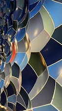 A close-up view of a mosaic pattern featuring an array of overlapping, curved shapes in various shades of blue and green, accented by a single red section. The design exhibits a smooth, reflective surface with soft highlights from lighting, creating a sense of depth and movement.