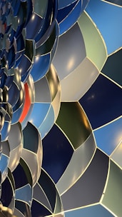 A close-up view of a mosaic pattern featuring an array of overlapping, curved shapes in various shades of blue and green, accented by a single red section. The design exhibits a smooth, reflective surface with soft highlights from lighting, creating a sense of depth and movement.