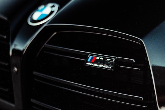 Close-up view of the front grille of a car with the BMW logo and M4 Competition badge visible. The design is sleek with dark tones and a shiny finish.