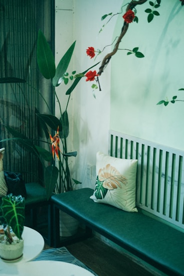 A cozy corner with a green bench adorned with a botanical-themed cushion. Vibrant red flowers hang from a wooden branch above, complemented by lush green leaves and a bird of paradise plant. A potted plant sits on a nearby round white table, adding to the natural decor.