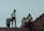 A group of people are gathered on a slightly inclined pavement. One person, dressed in a security uniform, stands near a woman who is bending over. Nearby, two men sit on a chair beside a small stand or cart, one in a striped shirt. The background is a muted sky, creating a silhouette effect of the figures.