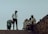 A group of people are gathered on a slightly inclined pavement. One person, dressed in a security uniform, stands near a woman who is bending over. Nearby, two men sit on a chair beside a small stand or cart, one in a striped shirt. The background is a muted sky, creating a silhouette effect of the figures.