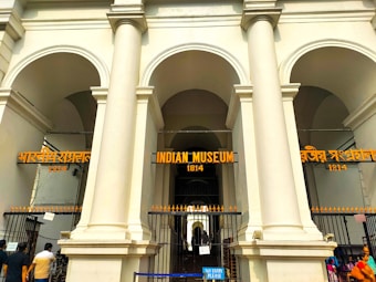 A grand entrance with large white pillars and arches leading to the Indian Museum, established in 1814. The museum's name is prominently displayed in three languages above the entrance. Several people are present near the gated entry, which is adorned with signs and a barrier.