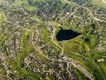 Aerial view of a residential area featuring numerous houses interspersed with greenery and winding roads, centered around a large lake. The layout displays an organized suburban development with clusters of homes and well-maintained lawns.