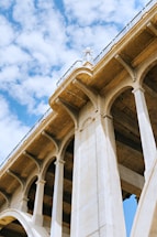 An architectural view featuring the underside of a large concrete bridge with multiple arches and columns. The bridge has ornate details and a vintage design. The sky is visible and filled with scattered white clouds, offering a contrast to the solid structure of the bridge.