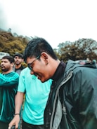 A group of young men are outdoors, dressed casually in sporty and weather-appropriate clothing. The main focus is a man wearing sunglasses, a black jacket, and smiling. Surrounding him are others in various poses, with greenery and trees in the background.