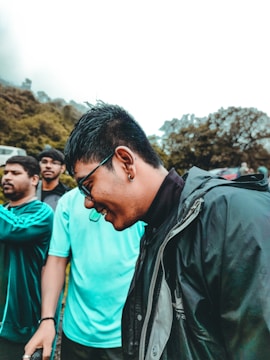 A group of young men are outdoors, dressed casually in sporty and weather-appropriate clothing. The main focus is a man wearing sunglasses, a black jacket, and smiling. Surrounding him are others in various poses, with greenery and trees in the background.