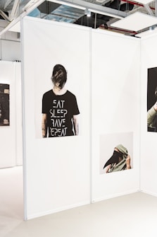 A gallery space with white partition walls displaying two different artworks. The first artwork shows a person with back turned, wearing a black t-shirt that reads 'EAT SLEEP RAVE REPEAT'. The second artwork features a person partially covering their face with a hood and a long, black beak-like mask.
