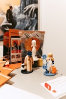 Several small figurines are arranged on a table, including a character holding a blue flower, a robed figure with a sheep near its feet, and a woman in white attire standing next to a stack of orange cylinders. Behind them, there is promotional material featuring various artistic designs and a larger image of a person with outstretched hands. The background also includes mountains and stylized birds in flight.
