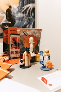Several small figurines are arranged on a table, including a character holding a blue flower, a robed figure with a sheep near its feet, and a woman in white attire standing next to a stack of orange cylinders. Behind them, there is promotional material featuring various artistic designs and a larger image of a person with outstretched hands. The background also includes mountains and stylized birds in flight.