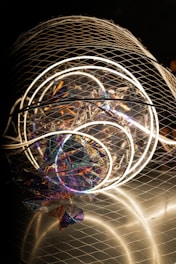A complex installation featuring illuminated circular neon lights enclosed in a wire mesh. The structure includes colorful, reflective materials creating a holographic effect, with various hues of purple, blue, and orange visible. The overall composition is abstract and artistic, capturing attention with both light and texture elements.