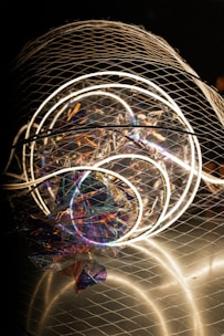 A complex installation featuring illuminated circular neon lights enclosed in a wire mesh. The structure includes colorful, reflective materials creating a holographic effect, with various hues of purple, blue, and orange visible. The overall composition is abstract and artistic, capturing attention with both light and texture elements.