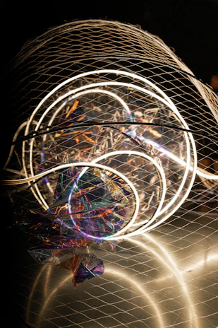 A complex installation featuring illuminated circular neon lights enclosed in a wire mesh. The structure includes colorful, reflective materials creating a holographic effect, with various hues of purple, blue, and orange visible. The overall composition is abstract and artistic, capturing attention with both light and texture elements.