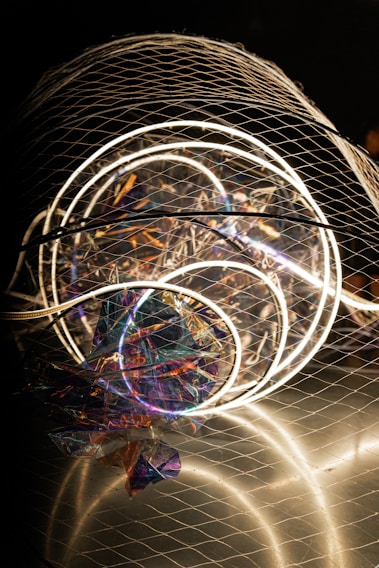 A complex installation featuring illuminated circular neon lights enclosed in a wire mesh. The structure includes colorful, reflective materials creating a holographic effect, with various hues of purple, blue, and orange visible. The overall composition is abstract and artistic, capturing attention with both light and texture elements.