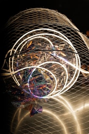A complex installation featuring illuminated circular neon lights enclosed in a wire mesh. The structure includes colorful, reflective materials creating a holographic effect, with various hues of purple, blue, and orange visible. The overall composition is abstract and artistic, capturing attention with both light and texture elements.