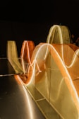 Curved structures illuminated by glowing, wavy lights are set against a dark background. These mesh structures in orange and beige hues create a dynamic visual effect. The scene is minimalistic yet intricate, with a play of light and shadow.
