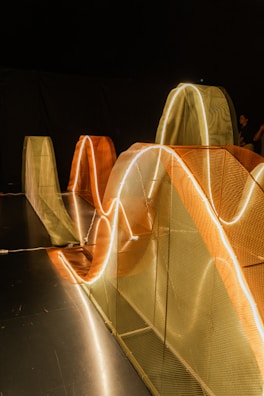Curved structures illuminated by glowing, wavy lights are set against a dark background. These mesh structures in orange and beige hues create a dynamic visual effect. The scene is minimalistic yet intricate, with a play of light and shadow.