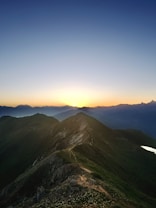 A breathtaking mountain landscape at sunset, with a rugged trail leading along the ridge. The sun is setting behind the distant peaks, creating a warm glow and casting long shadows. The sky transitions from deep blue at the top to a gentle gradient of orange near the horizon.
