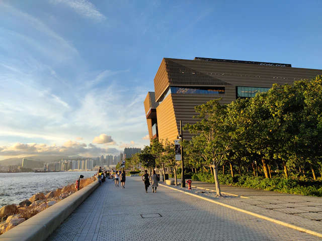 Waterfront promenade at West Kowloon Cultural District with the angular wood-clad M+ museum building at sunset.