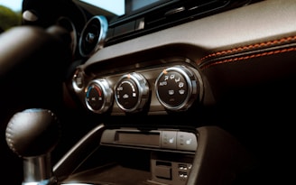 The interior of a car featuring the dashboard and air conditioning controls. The controls include dials for adjusting temperature and fan settings. The dashboard is detailed with black and brown materials, and noticeable orange stitching adds a touch of contrast.