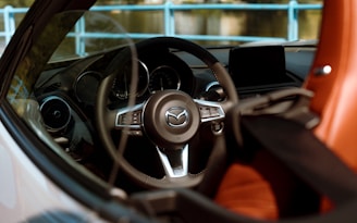 The interior of a car is visible through the driver's side window, showcasing a steering wheel with the Mazda logo at its center. The dashboard includes a modern display and various controls. The seat has a brown leather finish, and blue railings are faintly visible through the window, indicating an outdoor setting.