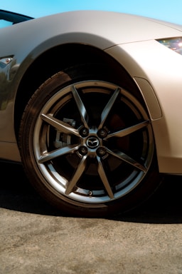 A close-up view of a car's shiny alloy wheel with the Mazda logo at the center. The car's body is a sleek metallic color, and the background features a clear blue sky. The sunlight creates reflections on the wheel and the car's fender, highlighting the smooth curves and design.