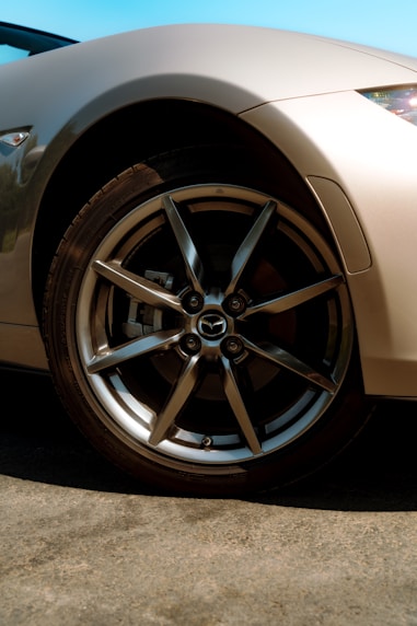 A close-up view of a car's shiny alloy wheel with the Mazda logo at the center. The car's body is a sleek metallic color, and the background features a clear blue sky. The sunlight creates reflections on the wheel and the car's fender, highlighting the smooth curves and design.