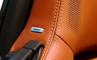 A close-up of a brown leather car seat or interior surface with detailed stitching. A BOSE metal emblem is embedded into the leather near some perforated holes, indicating a speaker system.
