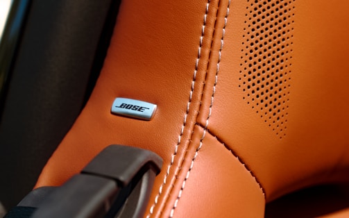 A close-up of a brown leather car seat or interior surface with detailed stitching. A BOSE metal emblem is embedded into the leather near some perforated holes, indicating a speaker system.
