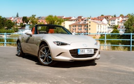 A sleek beige convertible car is parked on a road with a scenic backdrop of a river and a town with colorful buildings and greenery. The car is a Mazda MX-5 with shiny alloy wheels, and it features a Czech license plate. The sky is clear, indicating a bright sunny day.