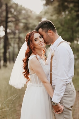 A couple embraces in a serene outdoor setting, with the woman wearing a white wedding dress and veil and the man dressed in a white shirt and suspenders. They appear to be in a forest with soft lighting, giving the scene a romantic and intimate atmosphere.