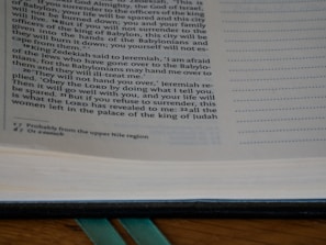 An open page of a book is shown, featuring text printed in a serif font. The text includes biblical references and explanations in a commentary style. The page number is visible at the bottom, and green ribbon bookmarks are partially seen at the spine.