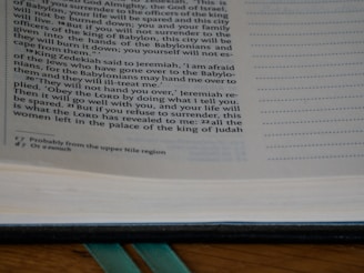 An open page of a book is shown, featuring text printed in a serif font. The text includes biblical references and explanations in a commentary style. The page number is visible at the bottom, and green ribbon bookmarks are partially seen at the spine.