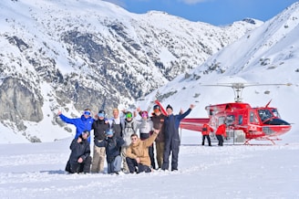 A group of people wearing winter clothing and ski goggles are posing together on a snowy mountain. In the background, a bright red helicopter with people standing near it is visible. The surrounding landscape features snow-covered peaks under a clear blue sky.